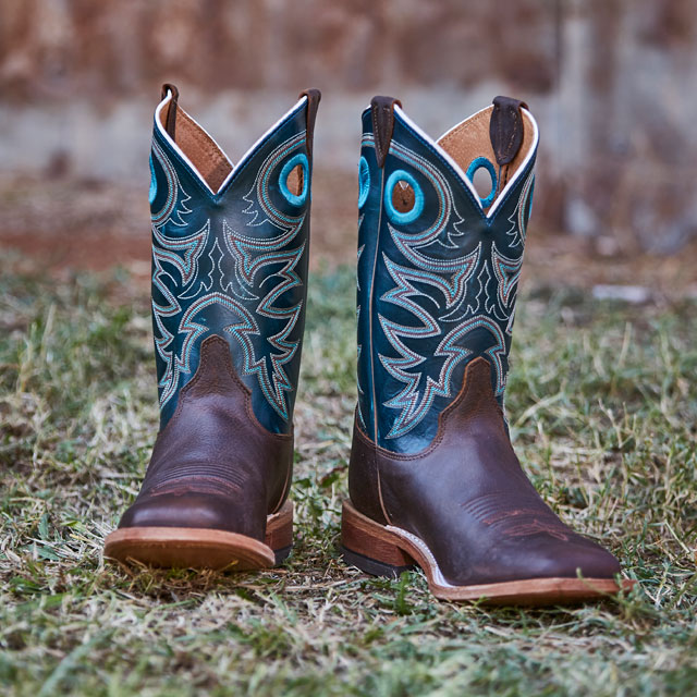 A pair of brown and blue men’s Justin Boots placed on grass with a tin background.